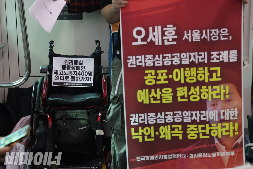 박 대표가 포체투지를 진행하는 동안 비어 있는 휠체어. 등받이에 “권리중심 중증장애인 해고노동자 400명 일터로 돌아가자!”라고 적힌 스티커가 붙어 있다. 그 옆에 한 활동가가 “오세훈 서울시장은 권리중심공공일자리 조례를 공포·이행하고 예산을 편성하라! 권리중심공공일자리에 대한 낙인·왜곡 중단하라!”라고 적힌 족자를 들고 있다. 사진 김소영